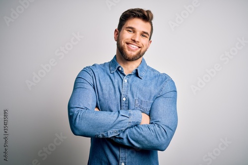 Young handsome blond man with beard and blue eyes wearing casual denim shirt happy face smiling with crossed arms looking at the camera. Positive person.