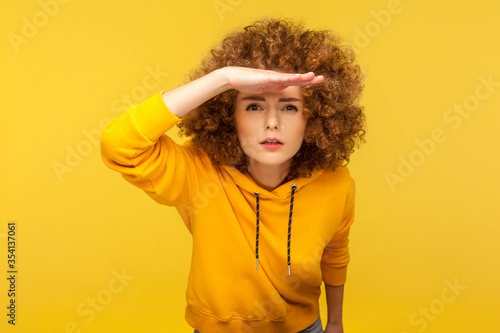 Portrait of curious curly-haired woman in urban style hoodie holding hand above eyes and peering into distance, looking far away, expecting and searching someone on horizon. studio shot isolated