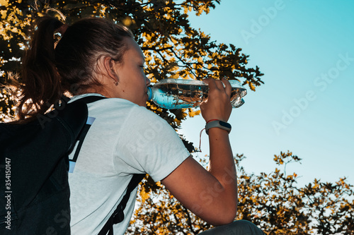 Young girl traveler with backpack quenches thirst from plastic bottle with cool mineral clear water. Energetic sports drink for water salt balance. Tired resting tourist, rear view. Hot sultry summer