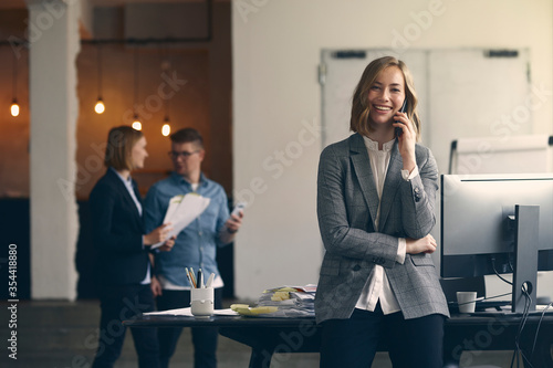 Portrait of smilling businesswoman talking on her phone in a modern work space, while her colleagues walks in the background
