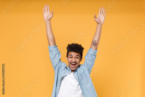 Joyful young african american guy in casual blue shirt posing isolated on yellow wall background studio portrait. People sincere emotions lifestyle concept. Mock up copy space. Spreading rising hands.