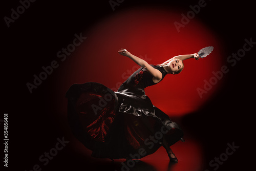 young flamenco dancer in dress holding fan while dancing on red and black