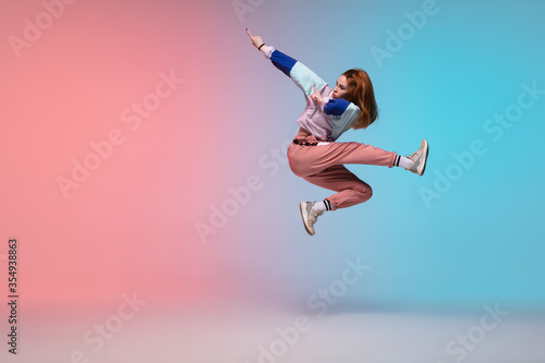 In jump. Beautiful girl dancing hip-hop in stylish clothes on colorful gradient background at dance hall in neon light. Youth culture, movement, style and fashion, action. Fashionable bright portrait.