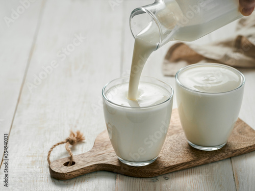 Pouring homemade kefir, buttermilk or yogurt with probiotics. Yogurt flowing from glass bottle on white wooden background. Probiotic cold fermented dairy drink. Trendy food and drink. Copy space left