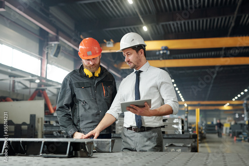 Industrial manager with tablet talking to worker and examining produced metal parts in factory shop