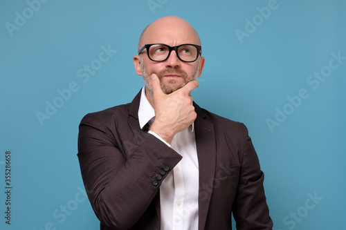 Thoughtful suspicious mature man looking up feeling skeptic doubtful. Businessman thinking holding hand on chin isolated on blue studio background