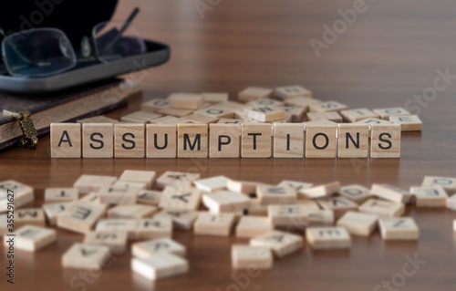 assumptions concept represented by wooden letter tiles on a wooden table with glasses and a book