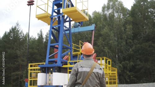 Oil field technical worker standing in front of crude oil pump unit. Engineer in orange helmet overseeing site of crude oil production. Extraction of oil.