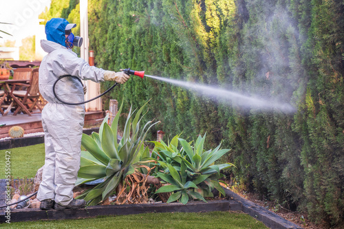 Fumigator applying plant protection products and herbicides to the plants of a garden house next to a Japanese-style stone fountain. The sprayer is wearing a protective mask and a white protective sui