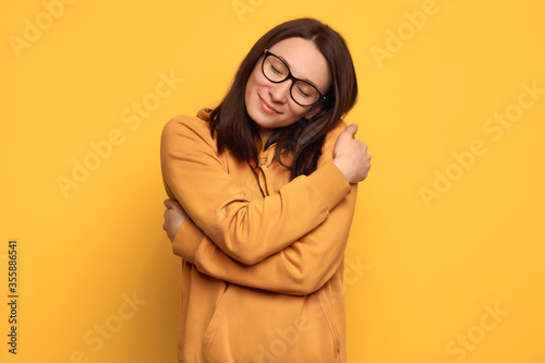 Closeup portrait confident smiling woman in glasses and orange cozy jumper holding and hugging herself isolated on yellow background. Positive human emotion. Love yourself and body positive concept