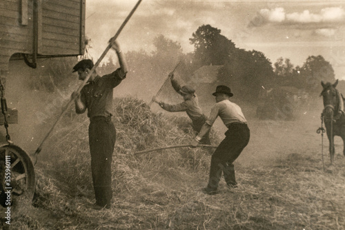 Latvia - CIRCA 1920s: Steam engine with thresher. Photo of farmers working on field. Archive vintage black and white photography