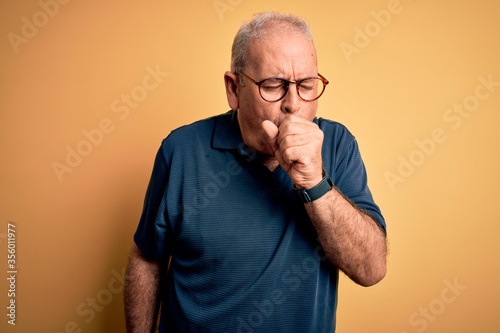 Middle age handsome hoary man wearing casual polo and glasses over yellow background feeling unwell and coughing as symptom for cold or bronchitis. Health care concept.