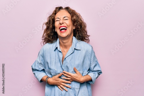 Middle age beautiful woman wearing casual denim shirt standing over pink background smiling and laughing hard out loud because funny crazy joke with hands on body.