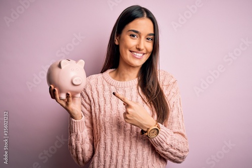 Young beautiful brunette woman holding piggy bank saving money for retirement very happy pointing with hand and finger