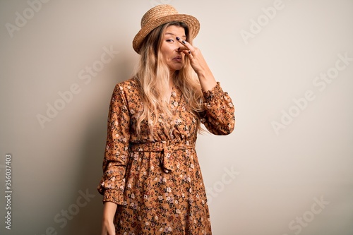 Young beautiful blonde woman wearing summer dress and hat over isolated white background smelling something stinky and disgusting, intolerable smell, holding breath with fingers on nose. Bad smell