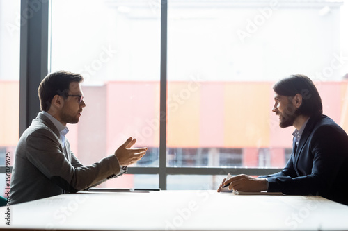 Side view two businessmen talking at negotiations solve current issues, sales manager make offer to client speaking sit at desk. Job interview process applicant and boss interaction in office concept