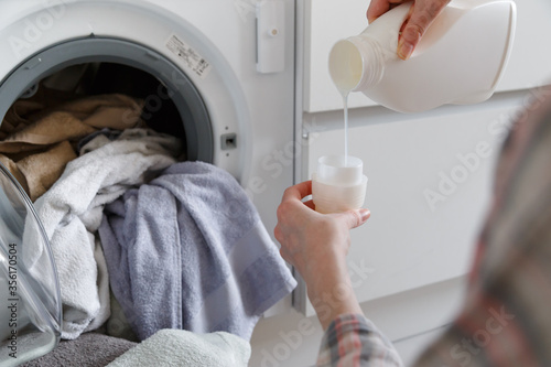 Close up of female hands pouring liquid laundry detergent into cap on white rustic table with towels on background in bathroom.