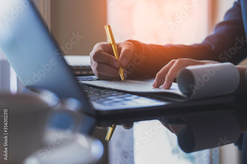 Business woman hand write and sign cheque book with laptop computer on the desk at office. Check payment, paycheck and payroll conceptual. Dark tone.