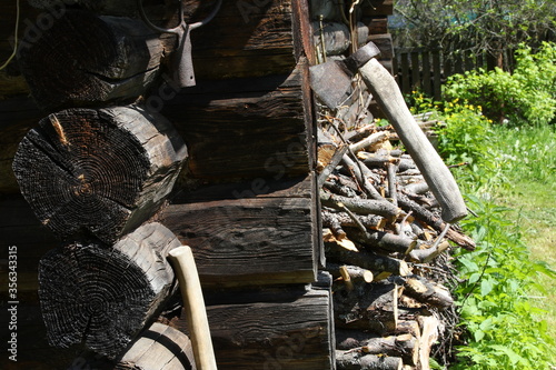 View of the corner of the house with a rusty axe in the wall and a woodpile on the plot with green grass. An element of village life.Russia