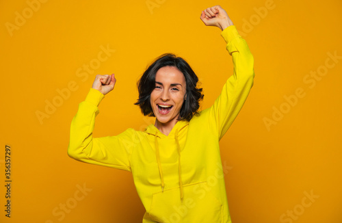 Yes! I've done it. Close-up photo of smiling satisfied girl dressed in a yellow hoodie, celebrating win with clenched fists, looking straight.