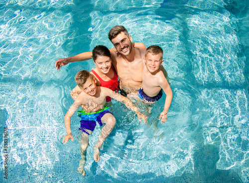 Top view of smiling family of four having fun and relaxing in indoor swimming pool at hotel resort.