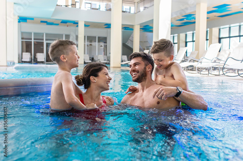 Smiling family of four having fun and relaxing in indoor swimming pool at hotel resort.