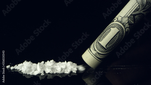 Line of cocaine being snorted up rolled up dollar bill on black reflective table top.