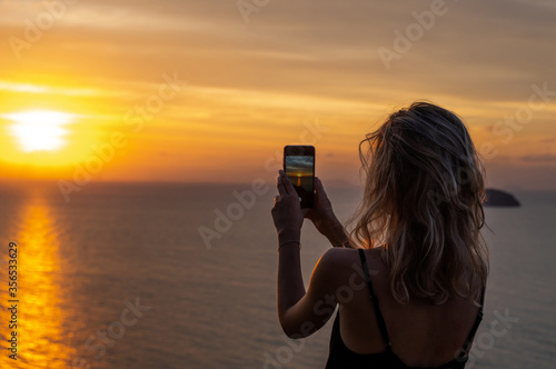 Woman hands holding mobile phone at sunset. Young curly hair woman taking photos with her cell phone in a beautiful amazing sunset over sea. Taking a picture on a smartphone during a vacation