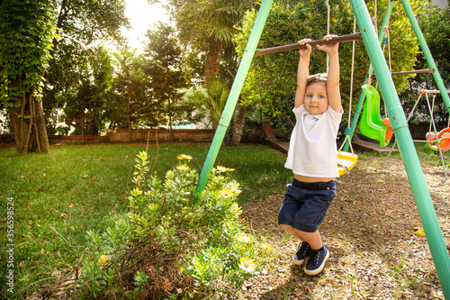 Bambino biondo con polo bianca si diverte vicino ad un altalena dentro un parco verde