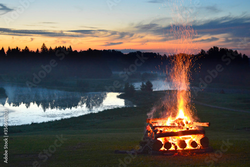 Midsummer national traditional celebration fire flames in the fireplace on grass field near river and forest with fog in sunset evening night sky clouds light