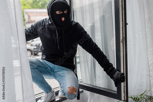 Selective focus of robber in balaclava standing on open window