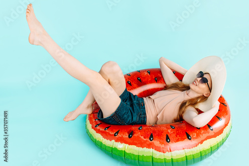 happy beautiful girl dressed in a swimsuit and a summer hat, sitting on an inflatable ring, resting isolated on a blue background