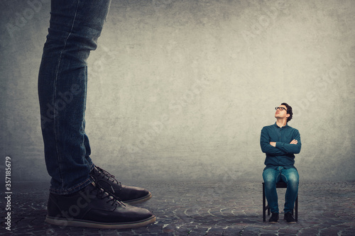 Tiny man employee seated on a chair looking up perplexed at his big boss. Office confrontation, motivation concept. Business person leadership concept.