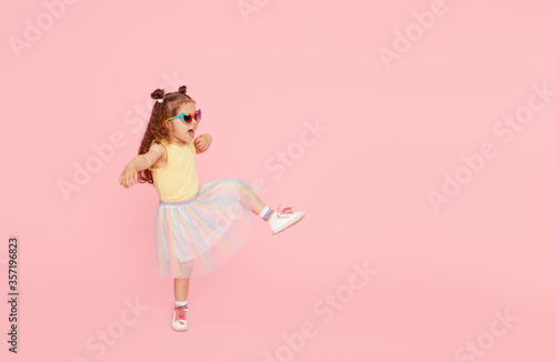 Portrait of surprised cute little toddler girl in sunglasses over pink background. Child model have fun and jump. Advertising childrens products