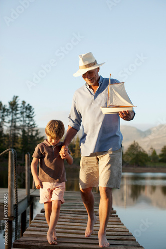 Grandfather grandson with toy sailboat walking along dock over lake