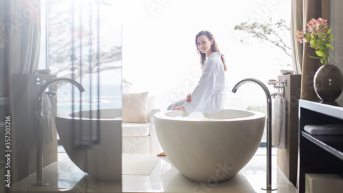 Portrait smiling woman wearing white bathrobe, sitting on edge of bathtub in bathroom