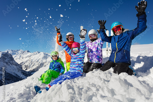 Ski school children class group portrait in colorful outfit throw snow in the air sitting together over mountain tops view and lift hands up