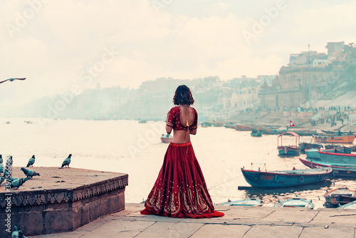 A beautiful Indian woman in a red Sari stands alone on the street. There is a flock of pigeons on the pedestal. In the background is a river and a view of the city. Travel and culture