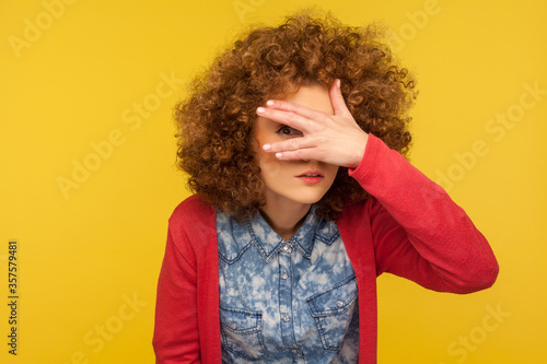 Portrait of curious nosy woman with curly hair spying rumors, looking through fingers with inquisitive expression, shy and scared to watch secret. indoor studio shot isolated on yellow background