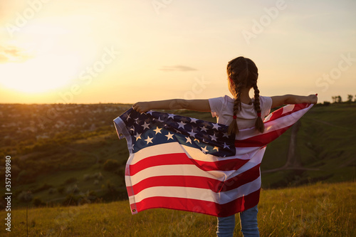 Independence Day USA. Rear view of a kid girl with the flag of America on nature at sunset.