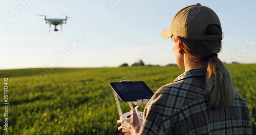 Close up of rear of Caucasian woman farmer in hat standing in green wheat field and controlling of drone which flying above margin. Female using tablet device as controller. Technologies in farming.