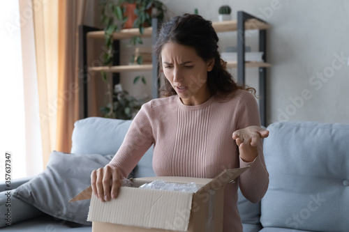 Angry confused woman unpacking parcel, wrong or broken online store order, sitting on couch at home, dissatisfied female looking in cardboard box, bad delivery service, displeased by post shipping