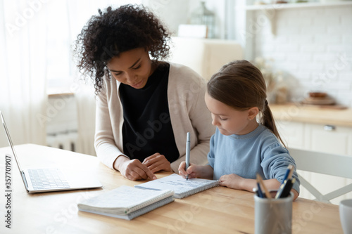 Home schooling conducted by parent or home tutor during quarantine time concept. African caring stepmom or nanny helps with school subject little daughter adorable kid girl, seated at table in kitchen