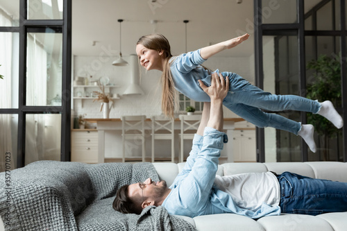 Happy family spend time together in living room father lying on couch lifts up little daughter, kid girl looking like plane imagines herself flying in air. Funny activities on weekend at home concept