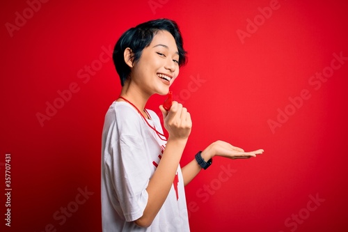 Young beautiful asian lifeguard girl wearing t-shirt with red cross using whistle pointing aside with hands open palms showing copy space, presenting advertisement smiling excited happy