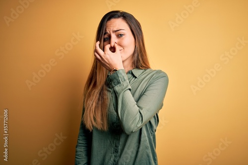 Young beautiful blonde woman with blue eyes wearing green shirt over yellow background smelling something stinky and disgusting, intolerable smell, holding breath with fingers on nose. Bad smell