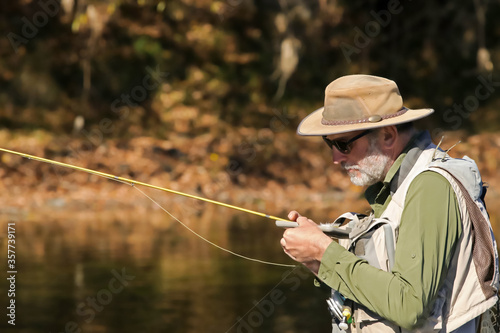 Mature bearded man choosing a fly from his fly box and holding his fishing rod amidst autumn colors
