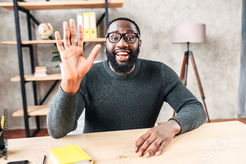 Excited African-American young guy in glasses looks into camera and greeting , waving hello. Video screen, video chat, online call, webcam shot