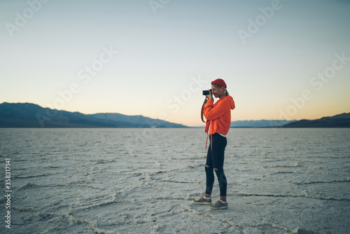 Young woman in touristic orange jacket making picture on camera during expedition to badwater national park, professional female photographer taking image of evening landscape in death valley