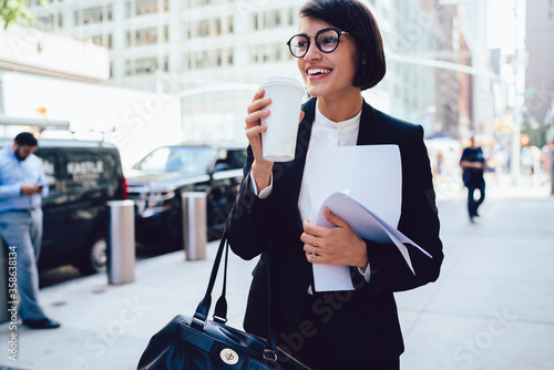 Cheerful female manager getting to work by foot drinking morning coffee to go while walking street, successful businesswoman in elegant wear enjoying sunny weather strolling with papers and documents